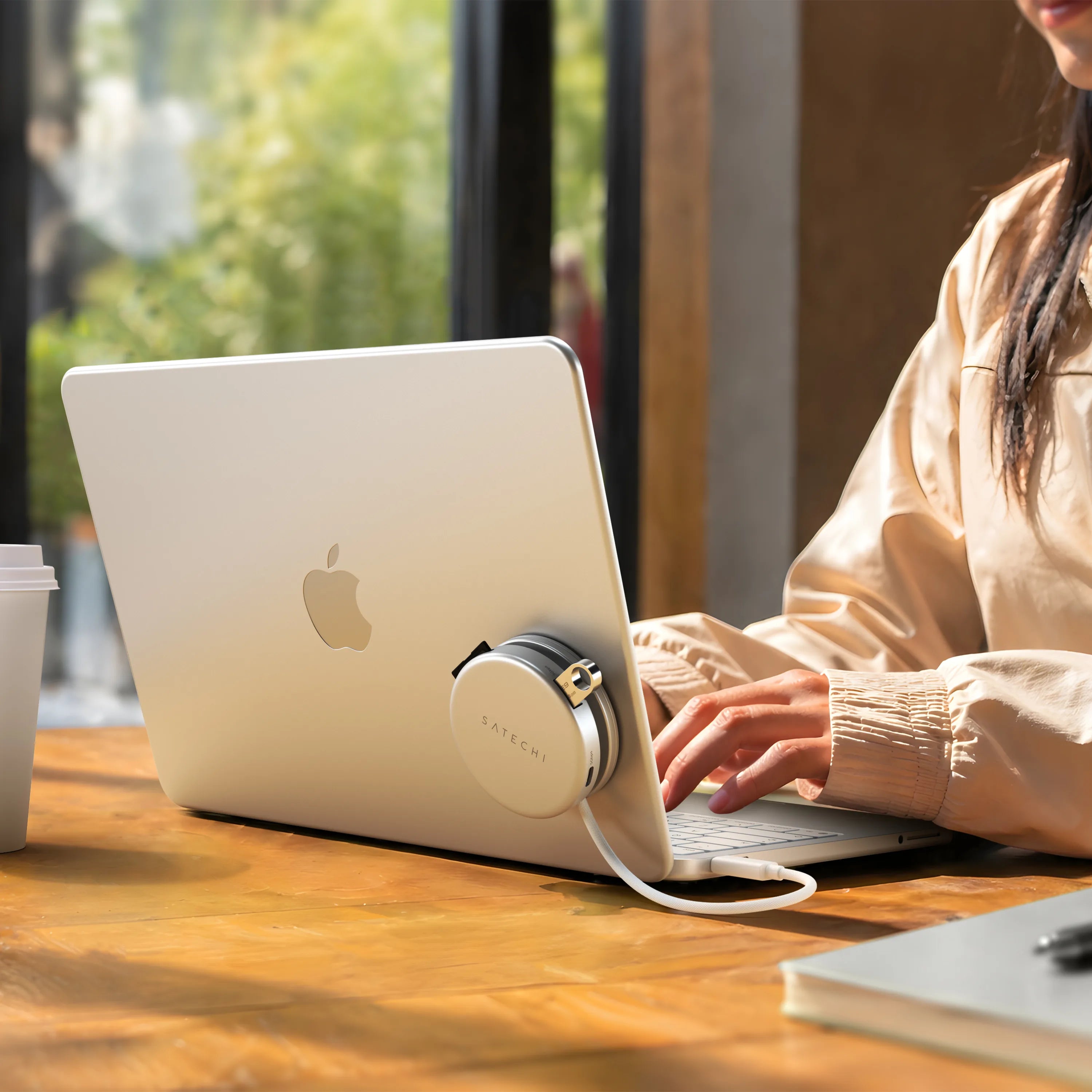 Silver Satechi OntheGo™ 5-in-1 Multiport Adapter magnetically mounted to the lid of a matching Citrus MacBook, with its color-matched braided USB-C cable plugged into the laptop, as a person types at a sunlit wooden table. The compact round magnetic USB-C hub for MacBook sits flush against the laptop's back panel.