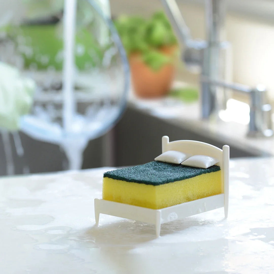 A Clean Dreams sponge, yellow and green, styled as a bed with pillows on a wet kitchen counter.