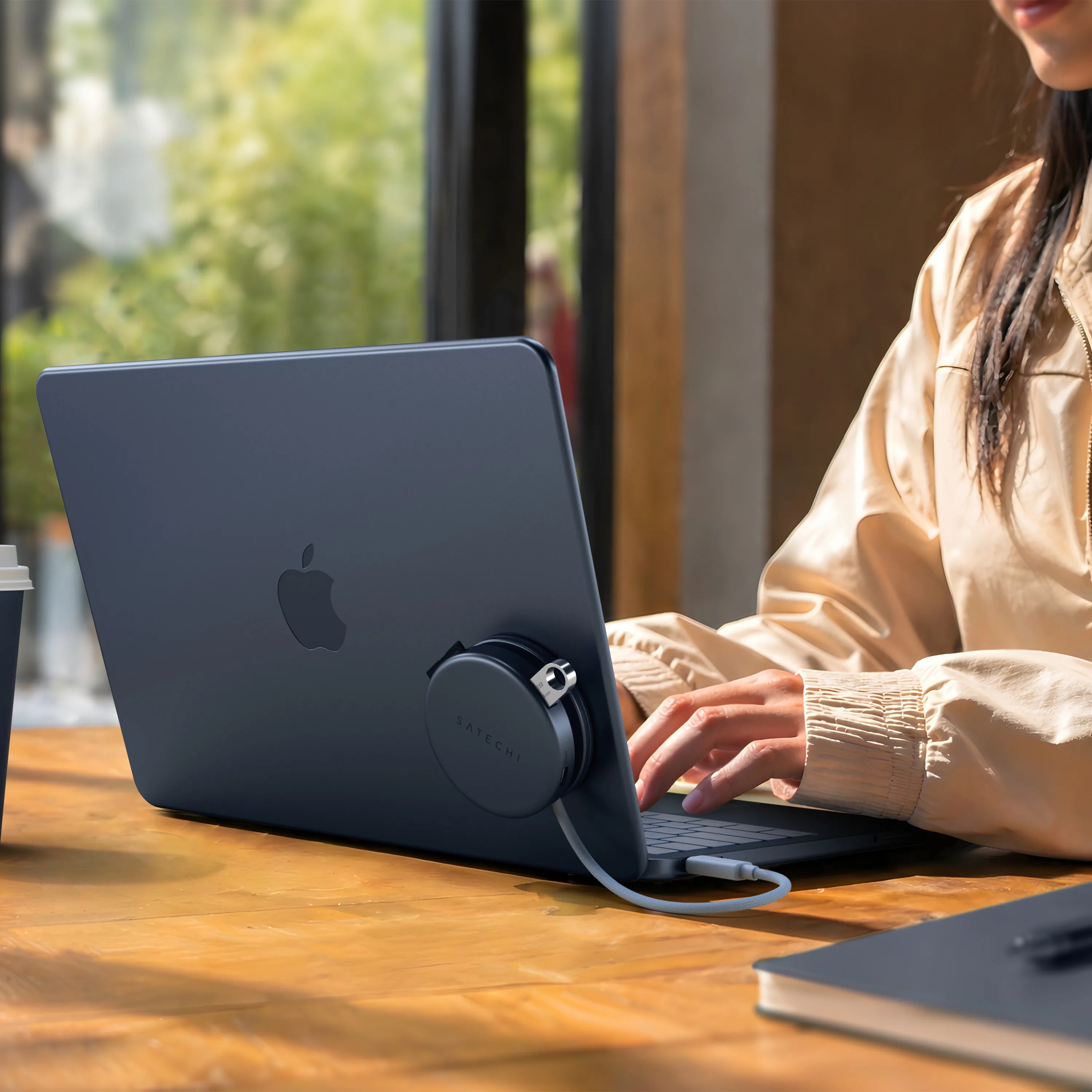 Indigo Satechi OntheGo™ 5-in-1 Multiport Adapter magnetically mounted to the lid of a matching Citrus MacBook, with its color-matched braided USB-C cable plugged into the laptop, as a person types at a sunlit wooden table. The compact round magnetic USB-C hub for MacBook sits flush against the laptop's back panel.