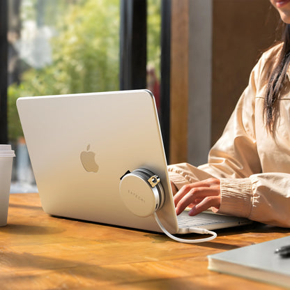 Silver Satechi OntheGo™ 5-in-1 Multiport Adapter magnetically mounted to the lid of a matching Citrus MacBook, with its color-matched braided USB-C cable plugged into the laptop, as a person types at a sunlit wooden table. The compact round magnetic USB-C hub for MacBook sits flush against the laptop's back panel.
