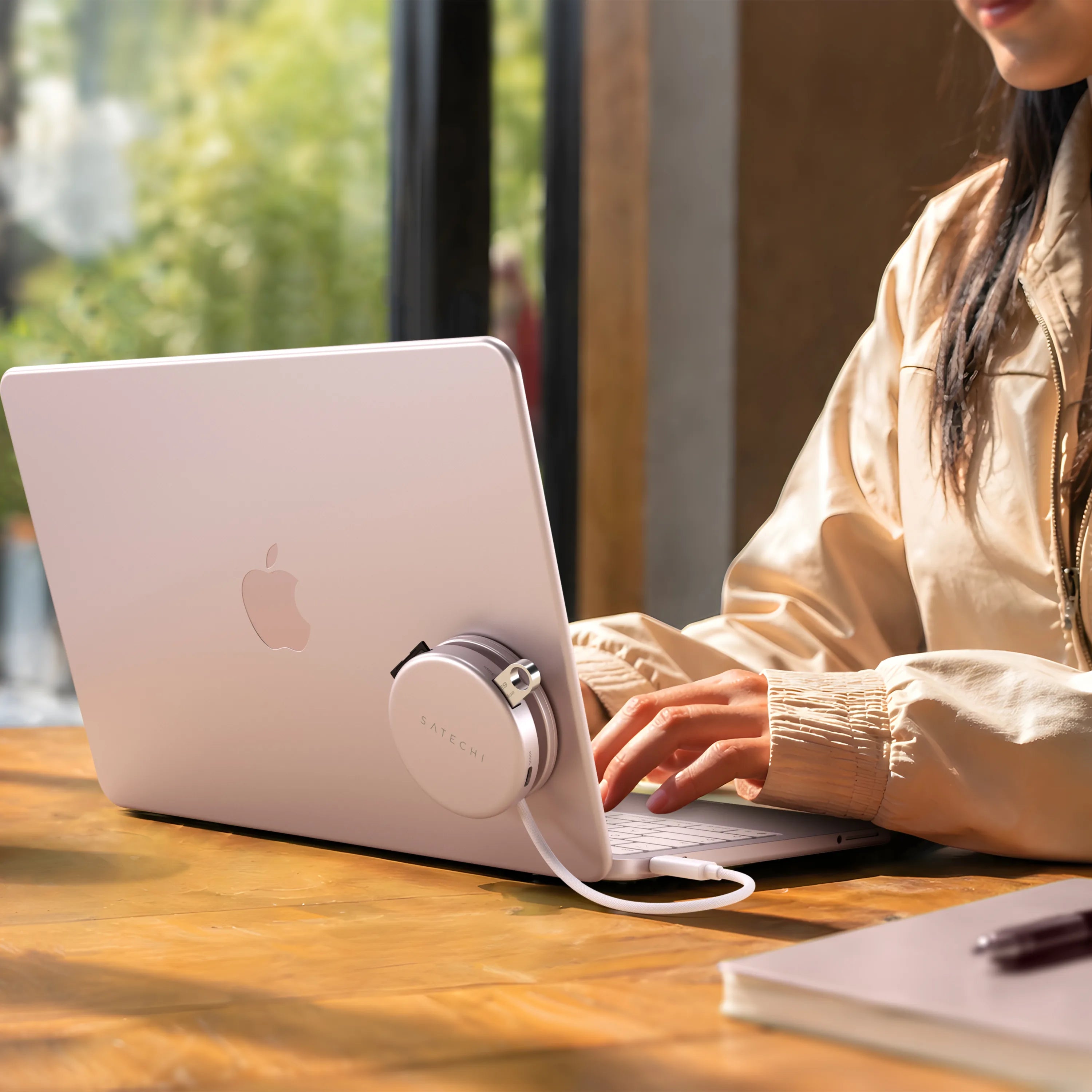 Blush Satechi OntheGo™ 5-in-1 Multiport Adapter magnetically mounted to the lid of a matching Citrus MacBook, with its color-matched braided USB-C cable plugged into the laptop, as a person types at a sunlit wooden table. The compact round magnetic USB-C hub for MacBook sits flush against the laptop's back panel.