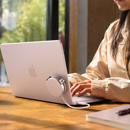 Blush Satechi OntheGo™ 5-in-1 Multiport Adapter magnetically mounted to the lid of a matching Citrus MacBook, with its color-matched braided USB-C cable plugged into the laptop, as a person types at a sunlit wooden table. The compact round magnetic USB-C hub for MacBook sits flush against the laptop's back panel.