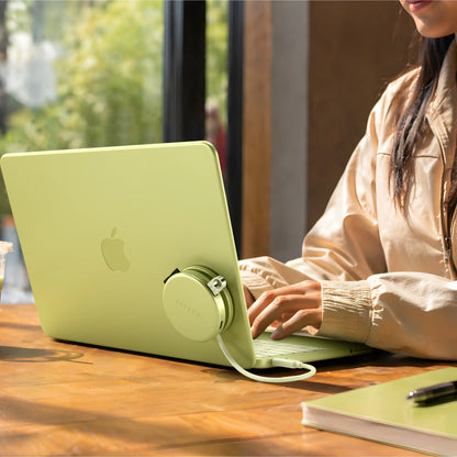 Citrus Satechi OntheGo™ 5-in-1 Multiport Adapter magnetically mounted to the lid of a matching Citrus MacBook, with its color-matched braided USB-C cable plugged into the laptop, as a person types at a sunlit wooden table. The compact round magnetic USB-C hub for MacBook sits flush against the laptop's back panel.