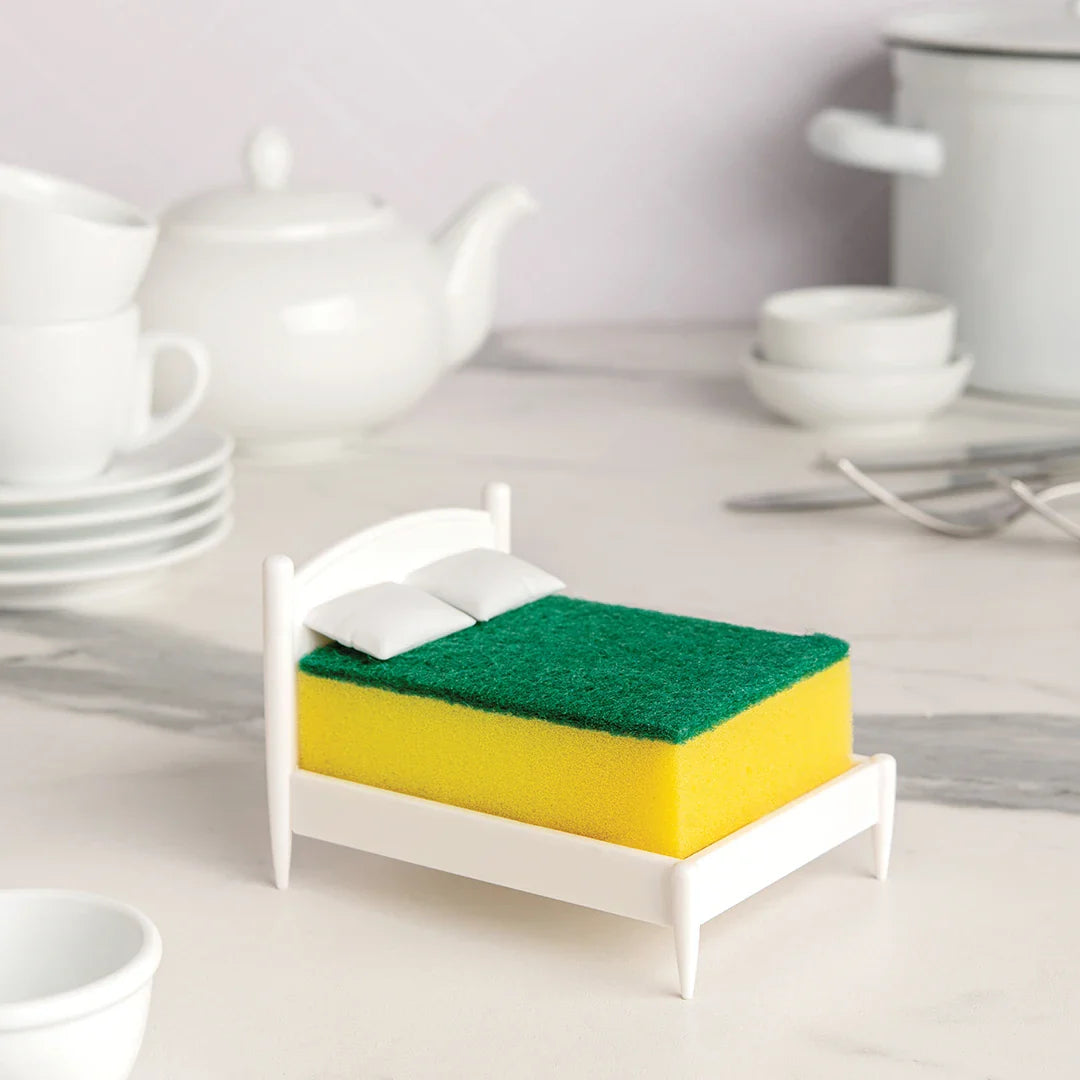 A scrub sponge rests on Clean Dreams with pillows, surrounded by OTOTO kitchenware on the counter.