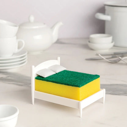 A scrub sponge rests on Clean Dreams with pillows, surrounded by OTOTO kitchenware on the counter.