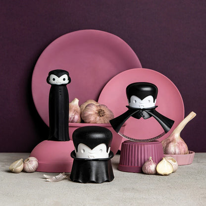 Three Drac N Roll kitchen tools with a garlic peeler and pink/purple plates in the background.